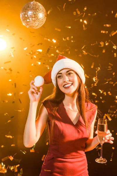 Schöne junge Frau mit Weihnachtsmütze und Champagnerglas unter fallendem Konfetti, Weihnachtskonzept — Stockfoto