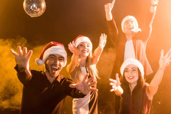 Group of friends in santa hats celebrating new year under yellow light — Stock Photo