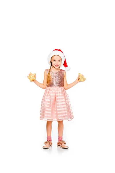 Lächelndes kleines Kind mit Weihnachtsmütze und Ingwerkeksen, das isoliert auf weiß in die Kamera blickt — Stockfoto