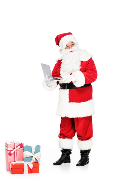 Santa claus com presentes segurando laptop e olhando para câmera isolada no branco — Fotografia de Stock