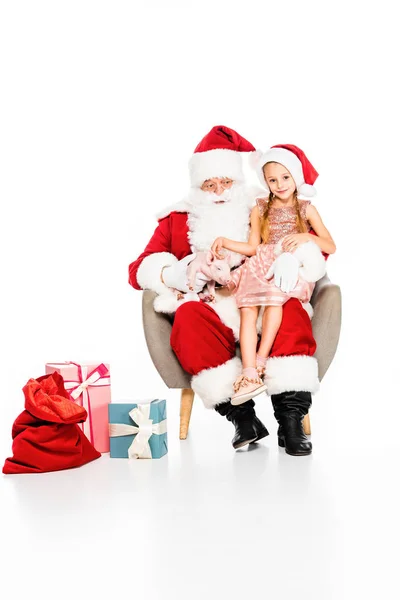 Santa claus y niño pequeño sentado en sillón con cerdito y mirando a la cámara aislada en blanco - foto de stock