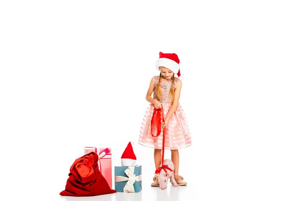 Adorable little child in santa hat with leashed piggy and pile of christmas gifts isolated on white — Stock Photo