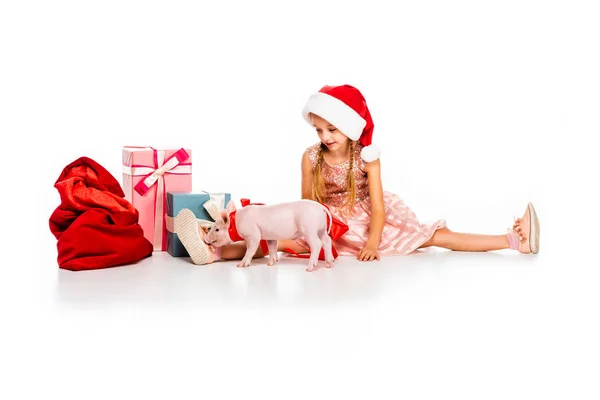 Adorable little child in santa hat with piggy and pile of christmas gifts isolated on white — Stock Photo