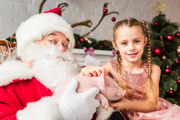 Weihnachtsmann und süßes kleines Kind spielen mit entzückendem Schwein und schauen in die Kamera — Stockfoto