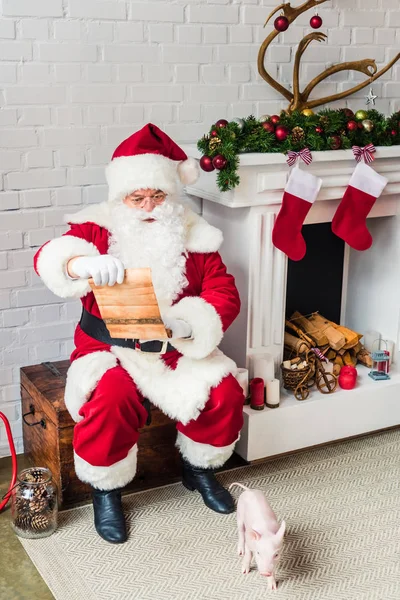 High angle view of santa claus sitting on chest near fireplace and reading wish list — Stock Photo