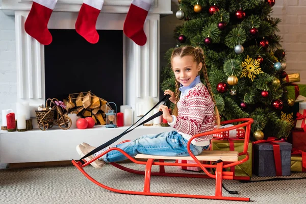 Carino bambino felice seduto sulla slitta e sorridente alla fotocamera vicino al camino — Foto stock