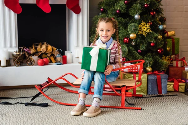 Entzückendes glückliches Kind sitzt auf Schlitten und hält Weihnachtsgeschenk in der Hand — Stockfoto