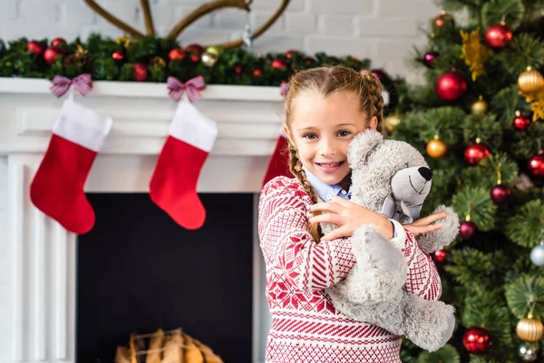 Adorable enfant heureux étreignant ours en peluche et souriant à la caméra au moment de Noël — Photo de stock
