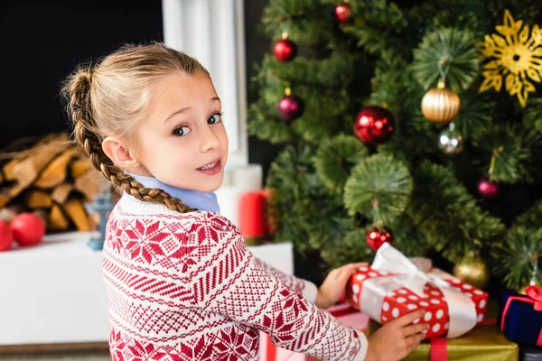 Criança adorável olhando para a câmera enquanto segurando presente sob a árvore de natal — Fotografia de Stock