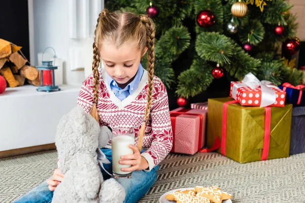 Adorable little child holding glass of milk and playing with teddy bear at christmas eve — Stock Photo