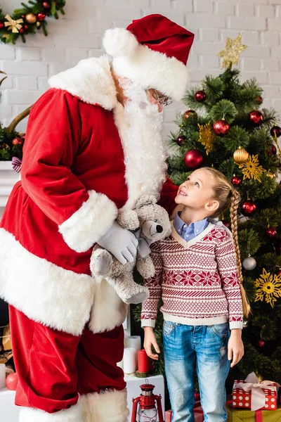 Санта держит плюшевого мишку и смотрит на милого улыбающегося ребенка, стоящего возле рождественской елки — стоковое фото