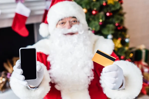 Санта Клаус держит кредитную карту и показывает смартфон с пустым экраном — стоковое фото