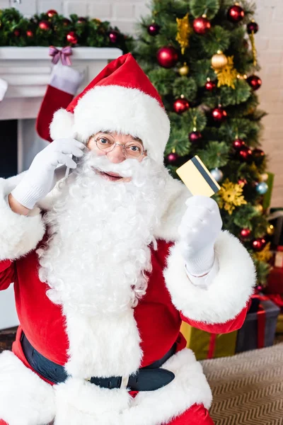 Weihnachtsmann hält Kreditkarte in der Hand und spricht per Smartphone — Stockfoto