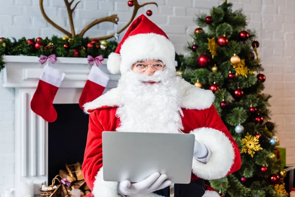 Санта Клаус с помощью ноутбука и смотреть на камеру возле камина — стоковое фото