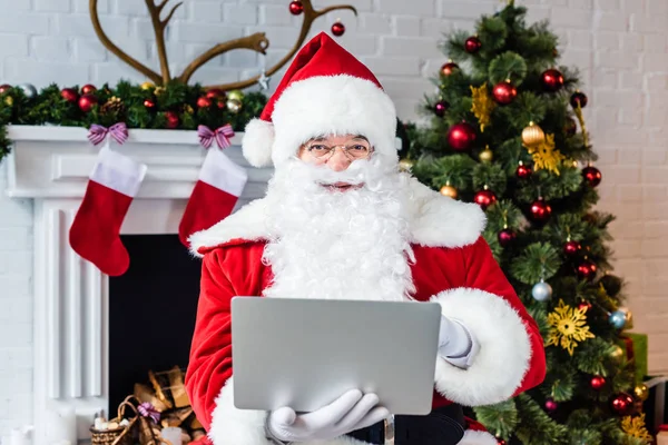 Santa claus usando laptop e olhando para a câmera perto da lareira — Fotografia de Stock