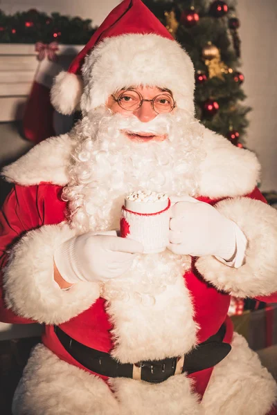 Père Noël tenant tasse avec boisson chaude et guimauves et regardant la caméra — Photo de stock