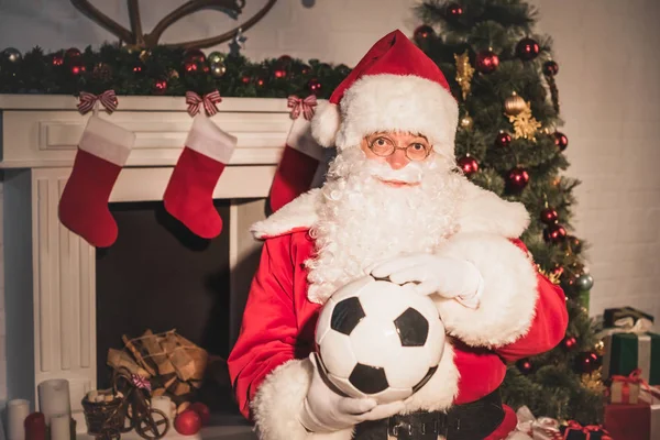 Санта Клаус тримає футбольний м'яч і дивиться на камеру, сидячи біля каміна — стокове фото