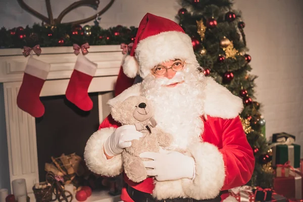 Santa claus sosteniendo oso de peluche y mirando a la cámara mientras está sentado cerca de la chimenea - foto de stock