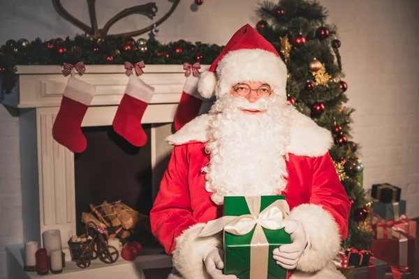 Père Noël claus tenant cadeau de Noël et regardant la caméra tout en étant assis près de la cheminée — Photo de stock