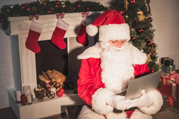 Vue grand angle de Santa Claus en utilisant un ordinateur portable près de la cheminée — Photo de stock