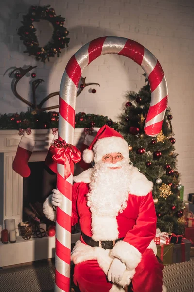 Санта тримає велику цукеркову тростину і дивиться на камеру біля каміна — стокове фото