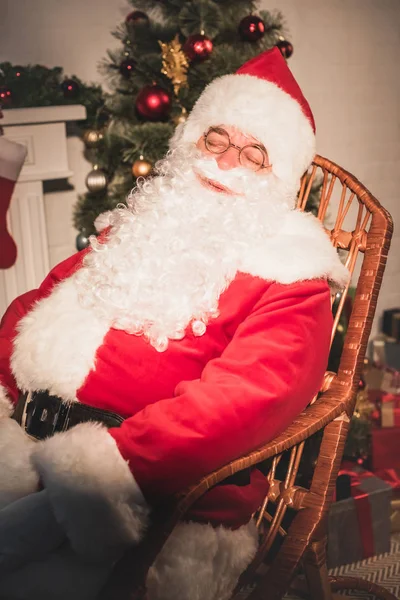 Санта Клаус спит в кресле-качалке возле камина и рождественской елки — стоковое фото