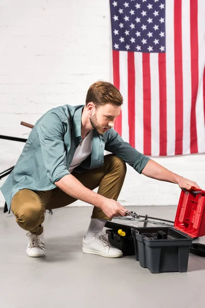 Joven guapo poniendo llave ajustable en caja de herramientas en frente de la pared con bandera americana - foto de stock