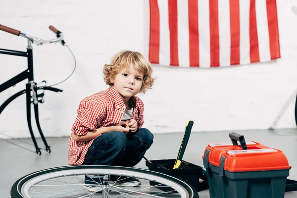 Adorable niño sentado cerca de herramientas cajas cerca de bicicleta desmontada en el taller — Stock Photo