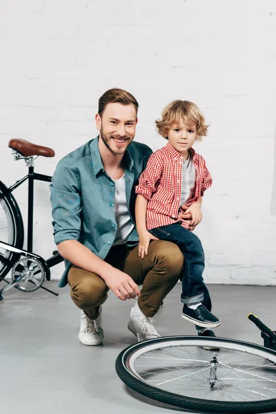 Счастливый мужчина сидит с маленьким сыном и смотрит на камеру в мастерской по изготовлению велосипедов — стоковое фото