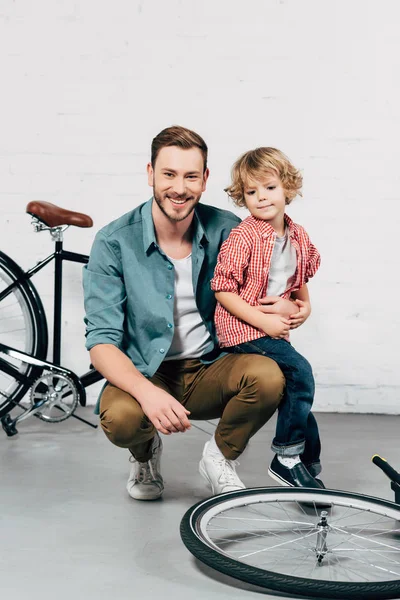 Joyeux bel homme assis avec petit fils et regardant la caméra dans l'atelier de vélo — Photo de stock