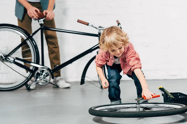 Joyeux petit garçon fixant roue par pinces tandis que son père debout derrière avec vélo démonté dans l'atelier — Photo de stock