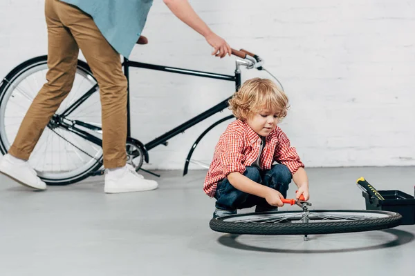 Foco seletivo da roda de fixação do menino por alicates, enquanto seu pai está atrás com a bicicleta desmontada na oficina — Fotografia de Stock