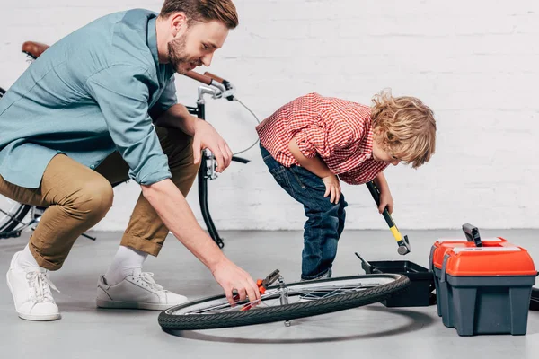 Боковой вид мужчины-ремонтника, который чинит велосипедное колесо плоскогубцами, в то время как его маленький сын стоит рядом с ящиком с инструментами в мастерской — стоковое фото