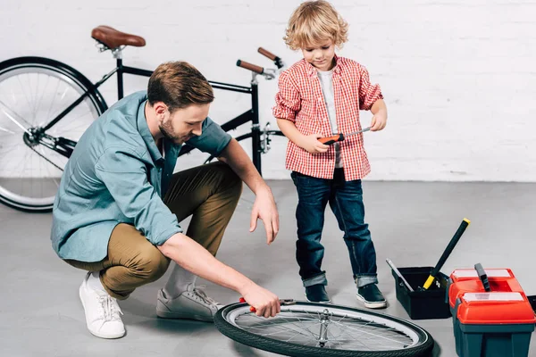 Riparatore maschio fissaggio ruota bicicletta con pinze mentre il suo piccolo figlio in piedi vicino in officina — Foto stock