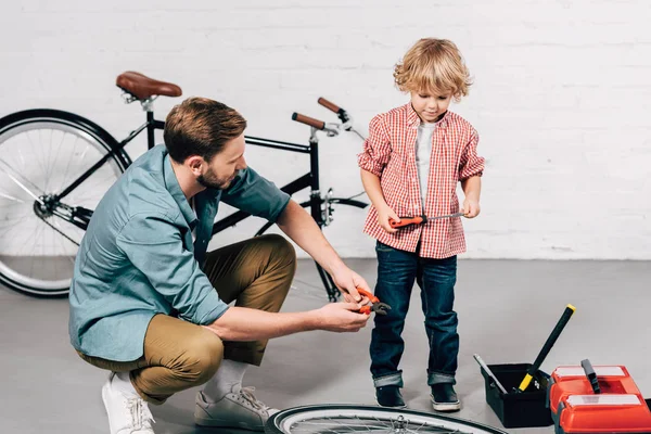 Вибірковий фокус ремонтанта фіксує велосипедне колесо з плоскогубцями, а його маленький син стоїть поруч з викруткою в майстерні — стокове фото