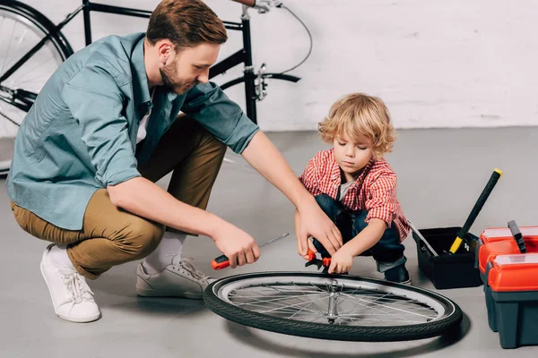 Sonriente hombre ayudando al pequeño hijo a fijar rueda de bicicleta con alicates en el taller — Stock Photo