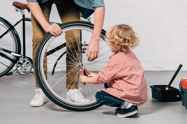 Cropped image of father holding bicycle wheel while his little son repairing with pliers at workshop — Stock Photo