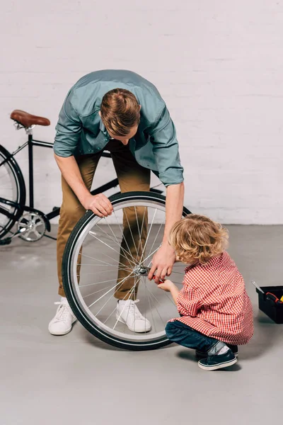 Homem ajudando filho pequeno reparar roda de bicicleta com alicate na oficina — Fotografia de Stock