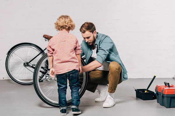 Hombre mostrando cómo reparar rueda de bicicleta a pequeño hijo en el taller - foto de stock