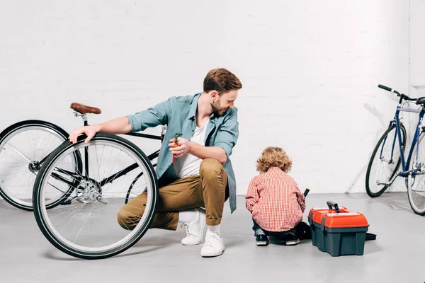 Cheerful male repairman holding screwdriver and looking at little son sitting near with tools boxes at workshop — Stock Photo