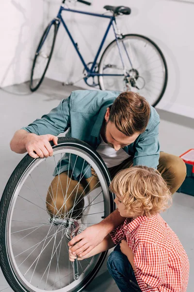 Vista de ángulo alto del reparador que muestra cómo fijar la rueda de bicicleta con llave ajustable al pequeño hijo en el taller - foto de stock