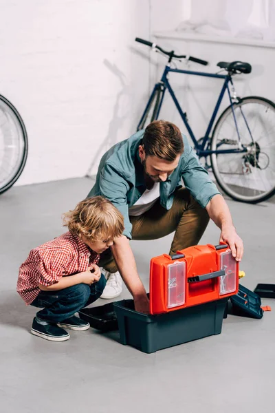 Высокий угол зрения отца и очаровательный маленький сын сидит рядом с открытым ящиком инструментов в велосипедной мастерской — стоковое фото