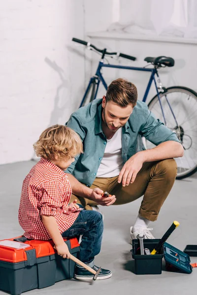 Hombre sosteniendo alicates y sentado con su pequeño hijo sentado cerca de las cajas de herramientas abiertas en el taller de bicicletas - foto de stock