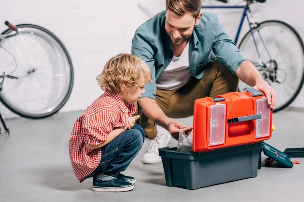 Pai e adorável filho sentado perto caixa de ferramentas abertas na oficina de bicicleta — Fotografia de Stock
