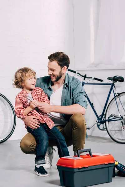 Uomo sorridente seduto con il piccolo figlio vicino alla cassetta degli attrezzi in officina per biciclette — Foto stock