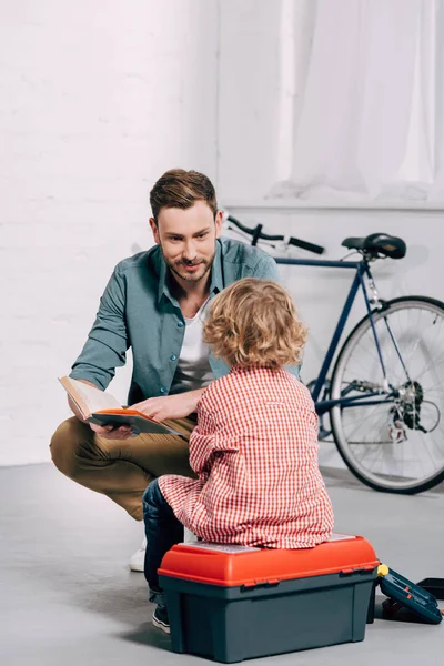 Hombre hablando y leyendo libro a pequeño hijo sentado en caja de herramientas en taller de bicicleta - foto de stock