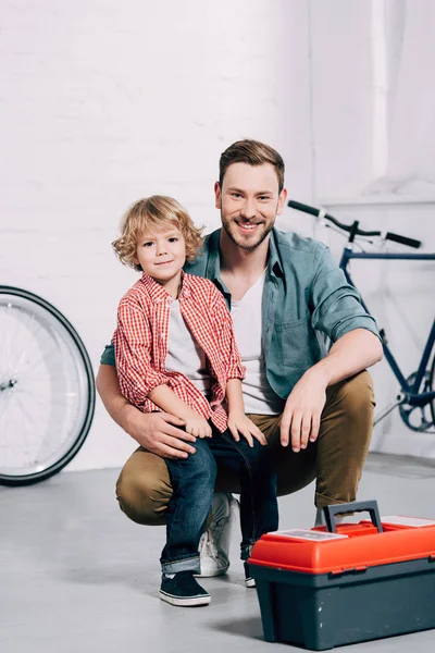 Felice uomo seduto con il piccolo figlio vicino cassetta degli attrezzi a officina di biciclette — Foto stock