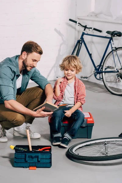 Hombre feliz señalando con la mano el libro al pequeño hijo sentado en la caja de herramientas en el taller de bicicleta - foto de stock