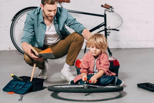 Selective focus of man with book sitting near little son fixing bicycle wheel with pliers in workshop — Stock Photo