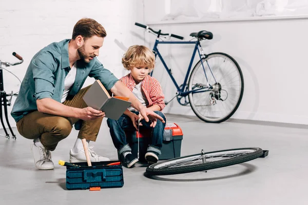 Uomo con libro che indica a mano la ruota della bicicletta al piccolo figlio in officina — Foto stock